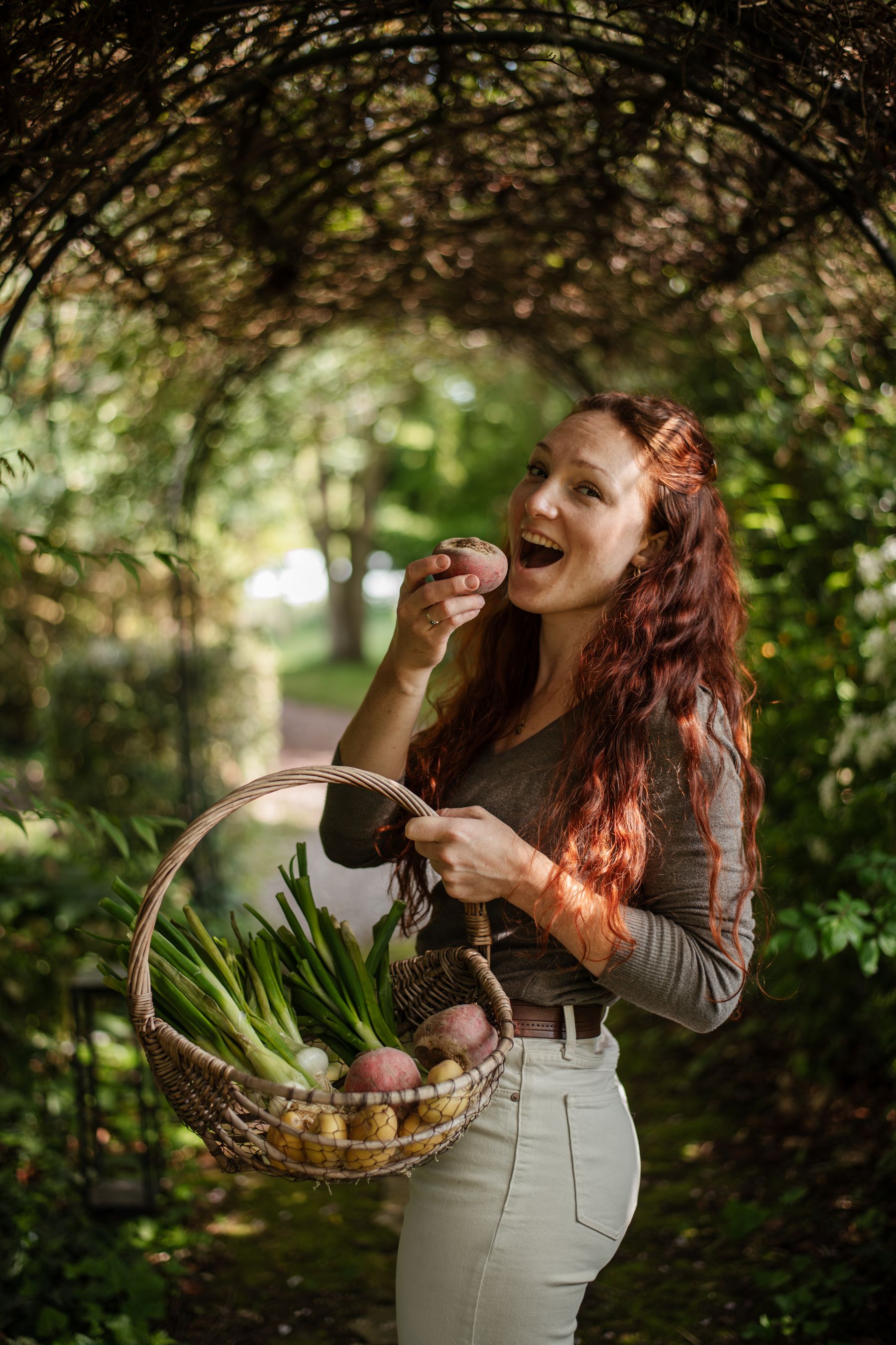 Laura Bridier riant sous une tonnelle de verdure, un panier de légumes au bras.