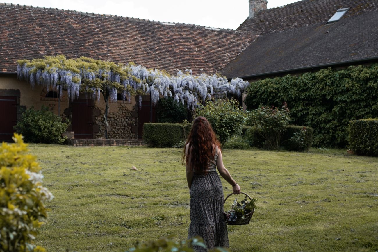 Laura Bridier traversant le jardin d'une longère couverte de glycine, un panier à la main.