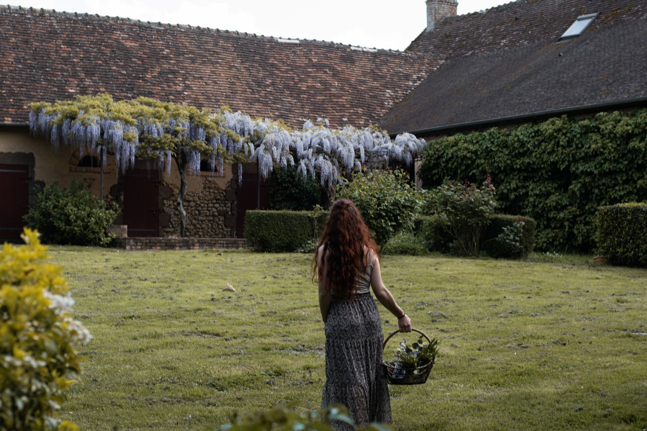 Laura Bridier traversant le jardin d'une longère couverte de glycine, un panier à la main.