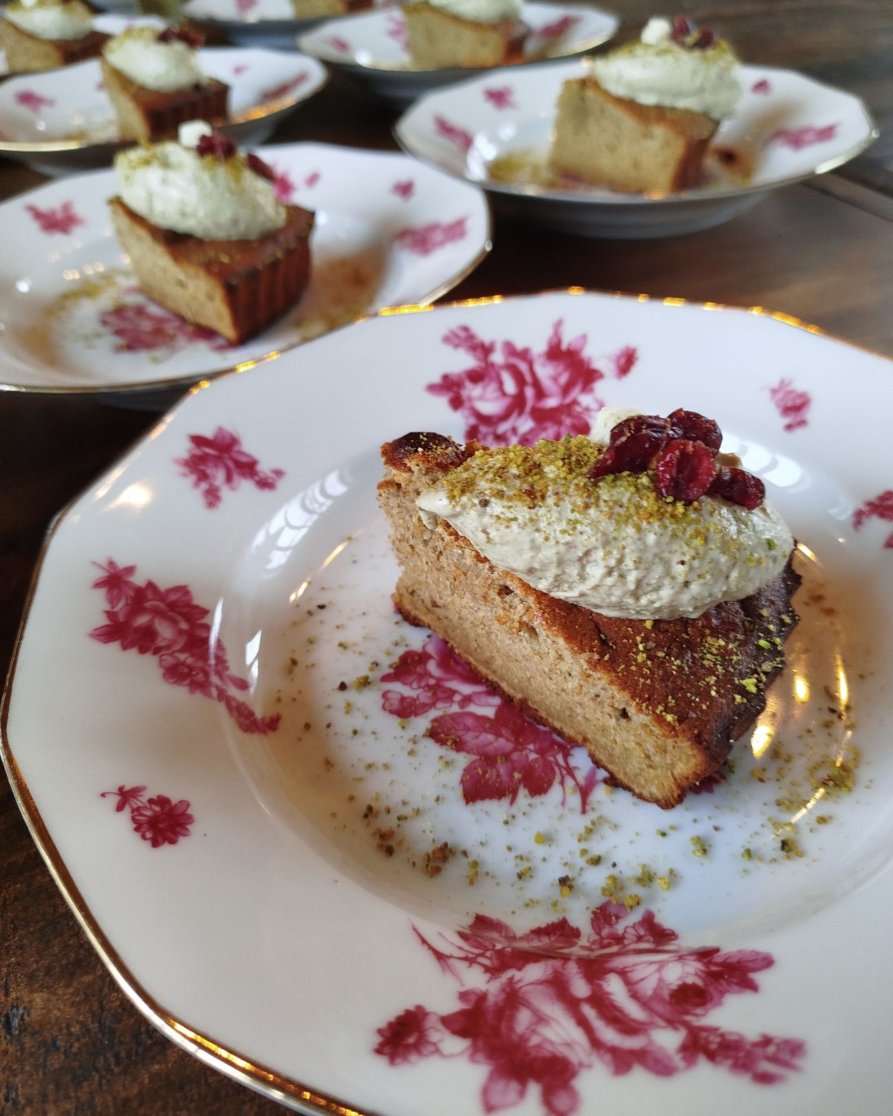 Parts de gâteau surmontées de crème, de pistaches et de fruits rouges, sur des assiettes à fleurs.