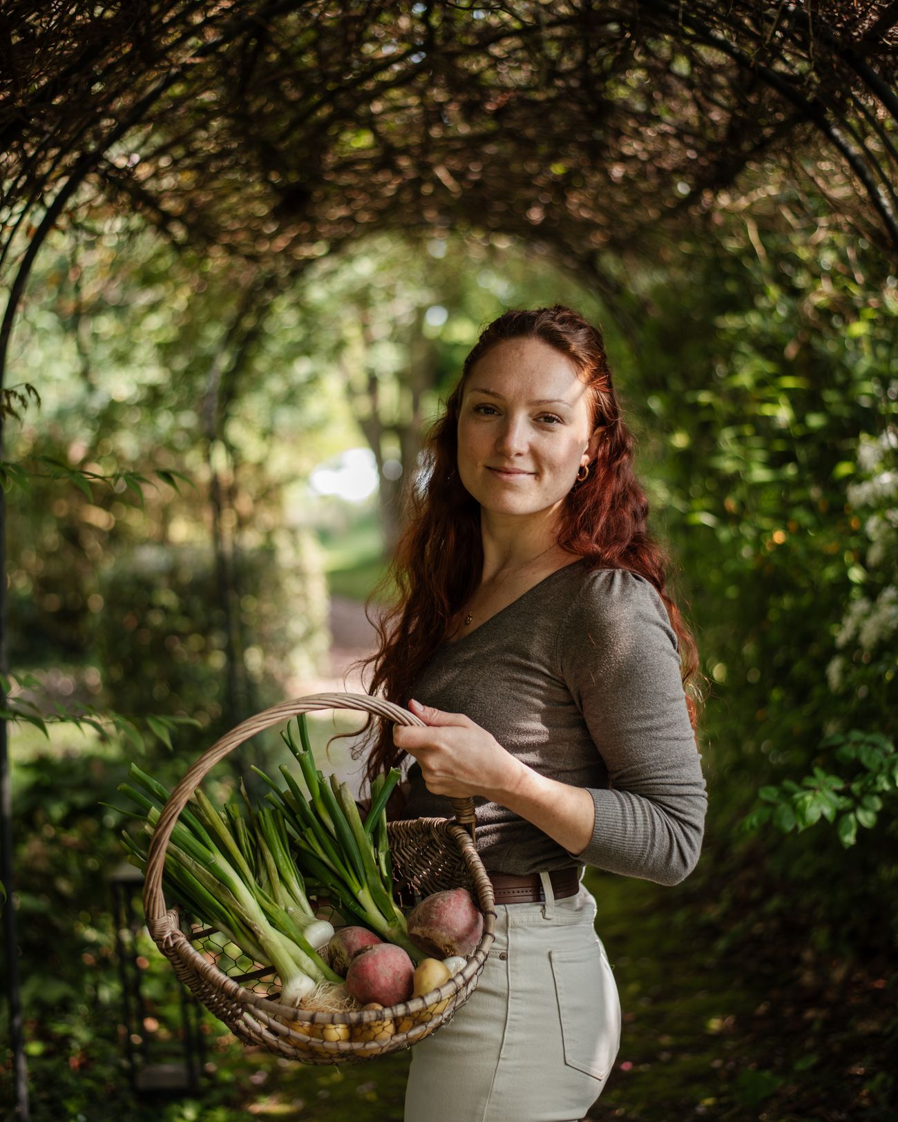 Portrait de Laura Bridier sous une tonnelle de verdure, un panier de légumes au bras.