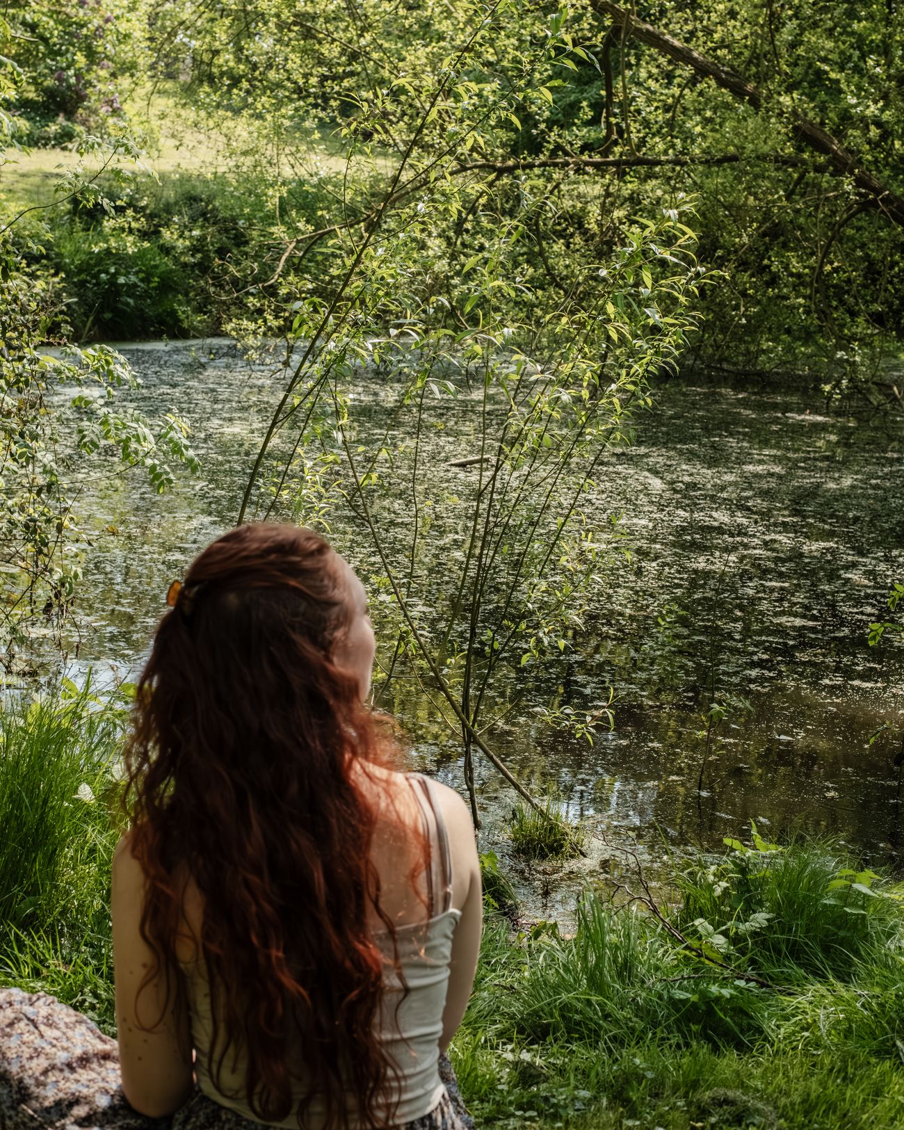 Laura Bridier de dos, assise au bord d'un étang, dans la verdure.